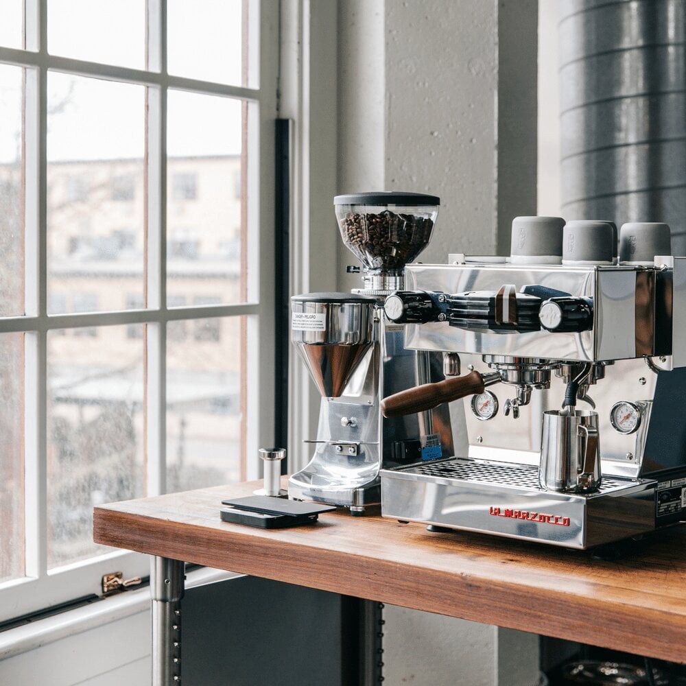 Espresso machine and grinder setup on wooden counter by large window in modern loft space