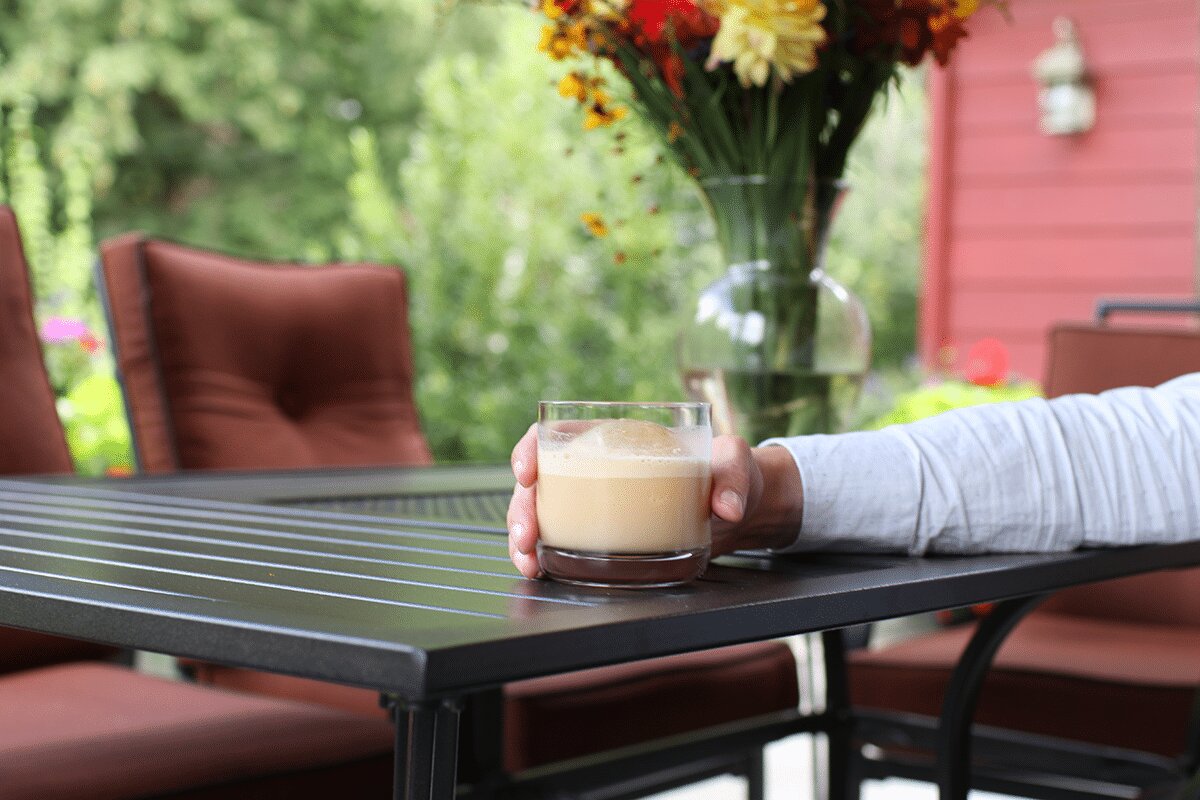 Person holding a cappuccino at an outdoor patio table with fresh flowers and a red chair in the background
