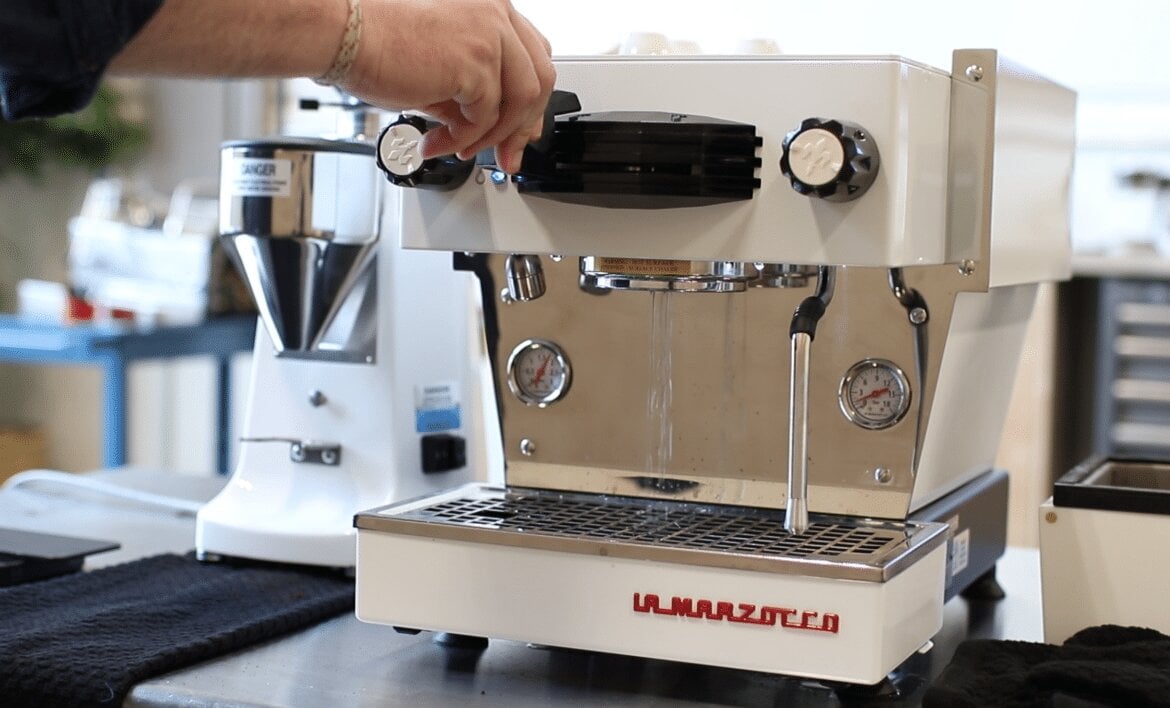 Barista pulling espresso shot on La Marzocco Linea Mini espresso machine in cream, with grinder and café equipment in background