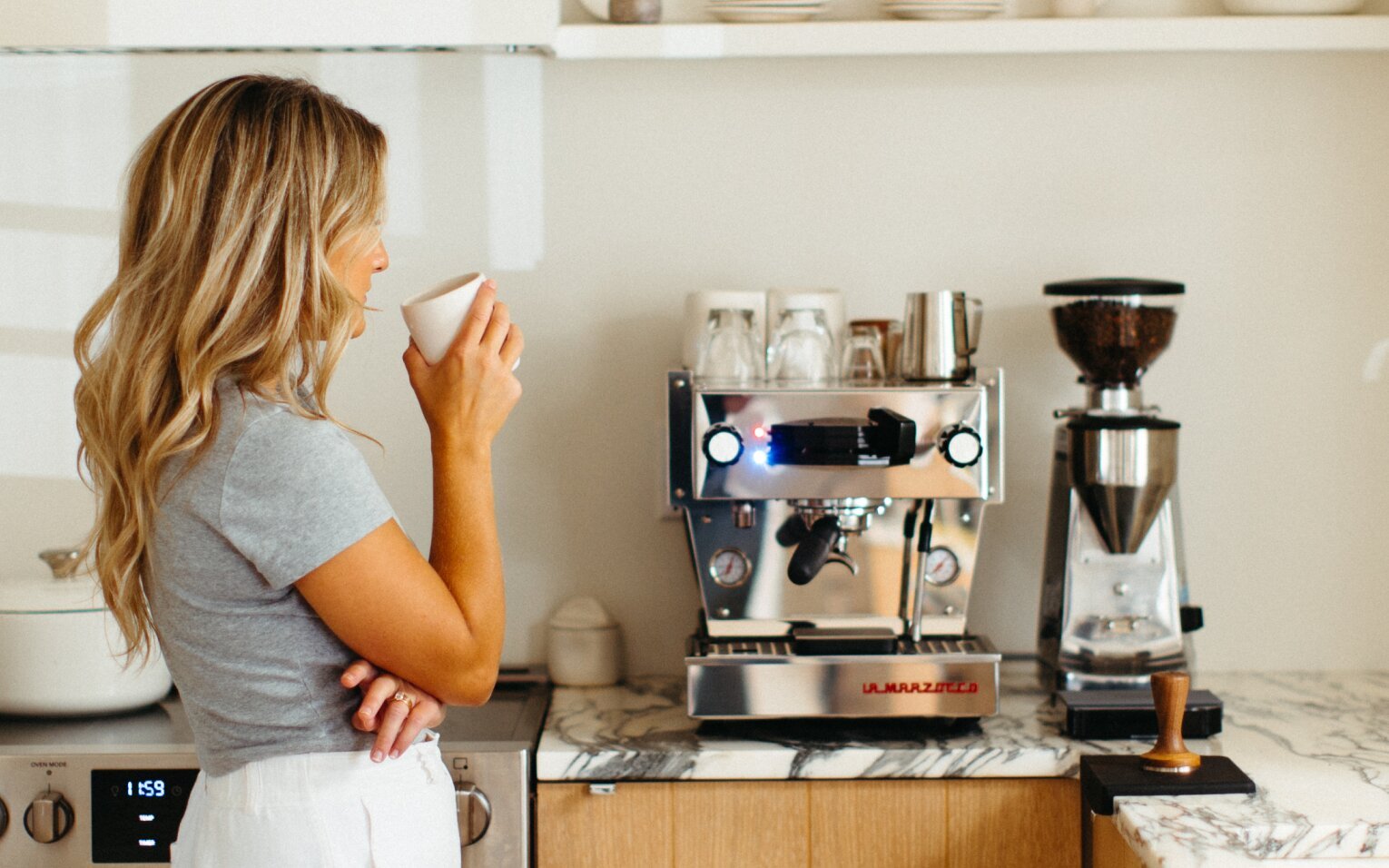 Woman holding a white cup in front of a La Marzocco espresso machine and grinder on a kitchen counter