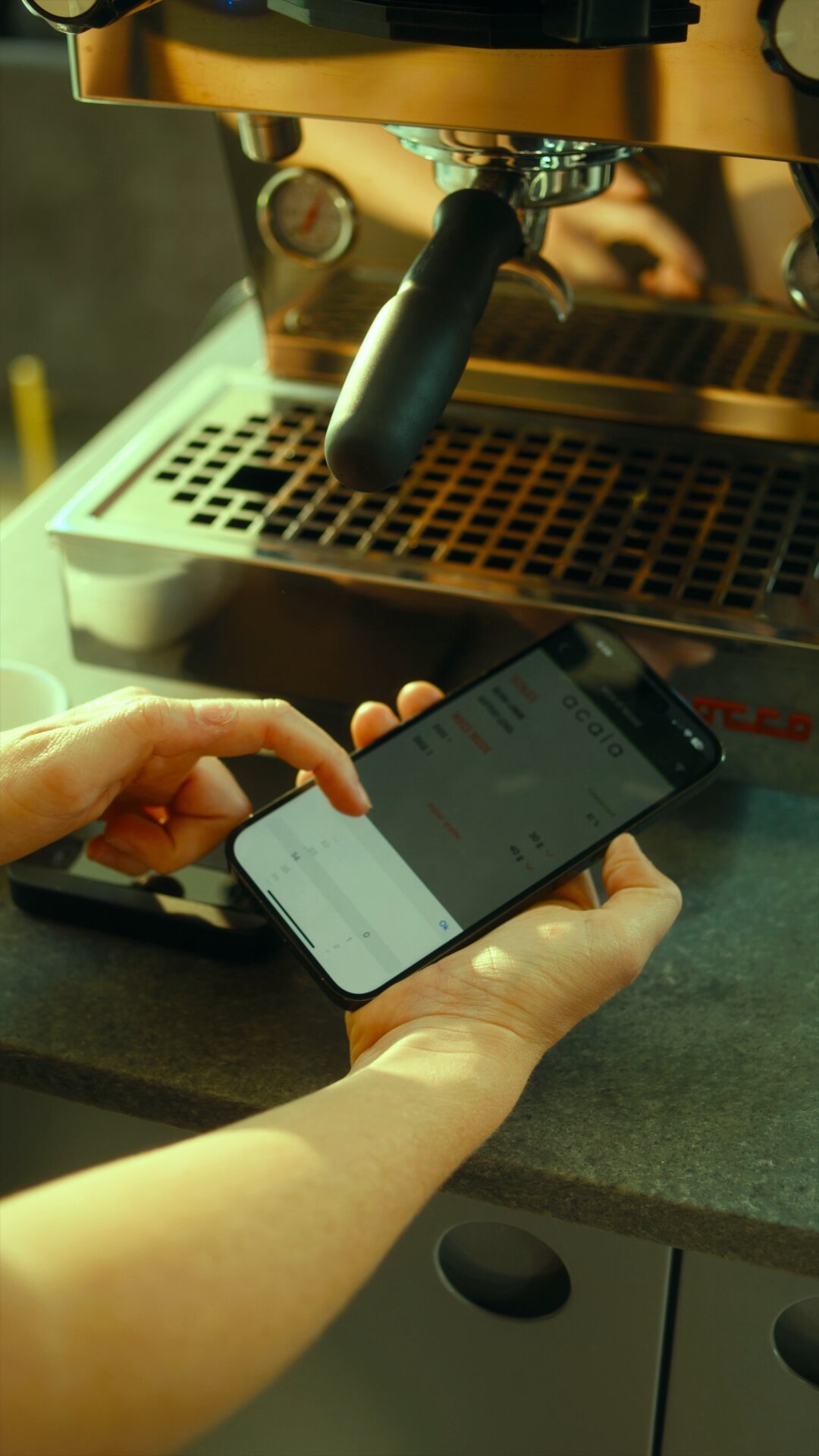 Home barista using a smartphone next to a La Marzocco espresso machine with portafilter and drip tray