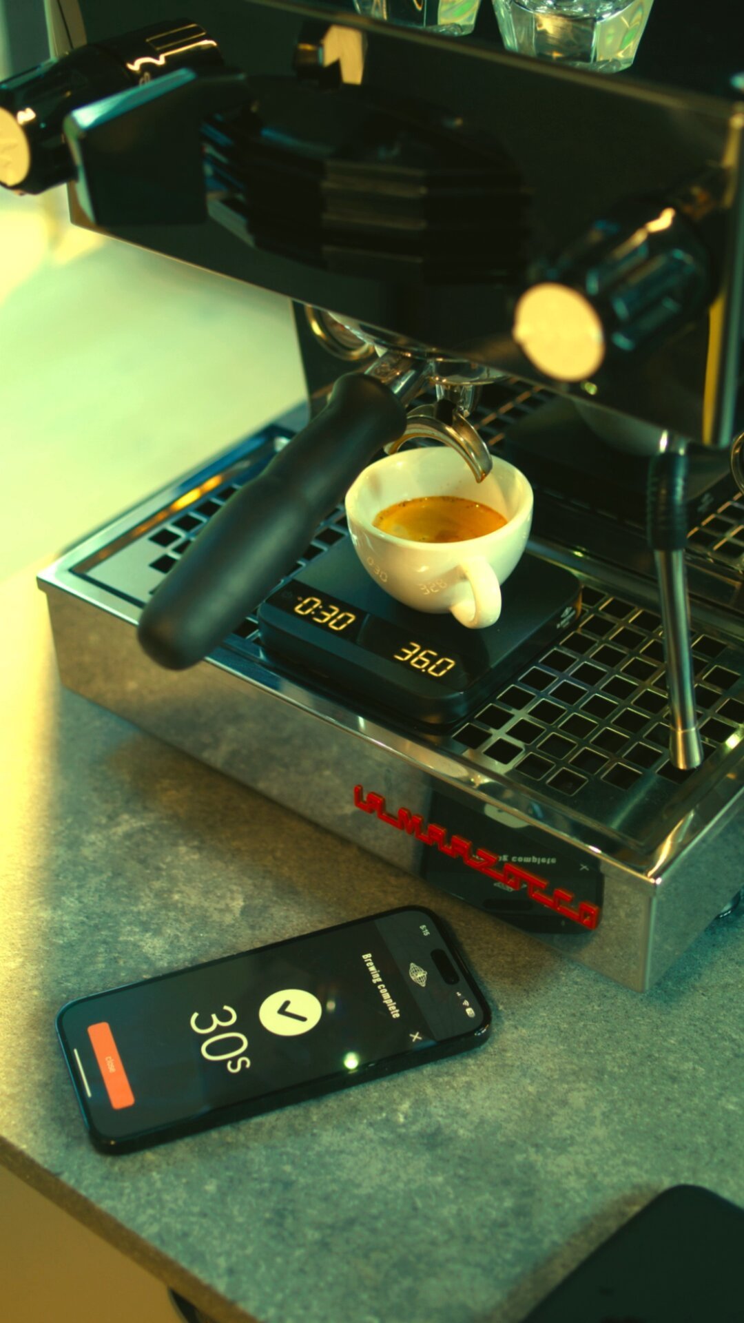 Espresso brewing on La Marzocco espresso machine with smartphone displaying temperature control, lifestyle scene