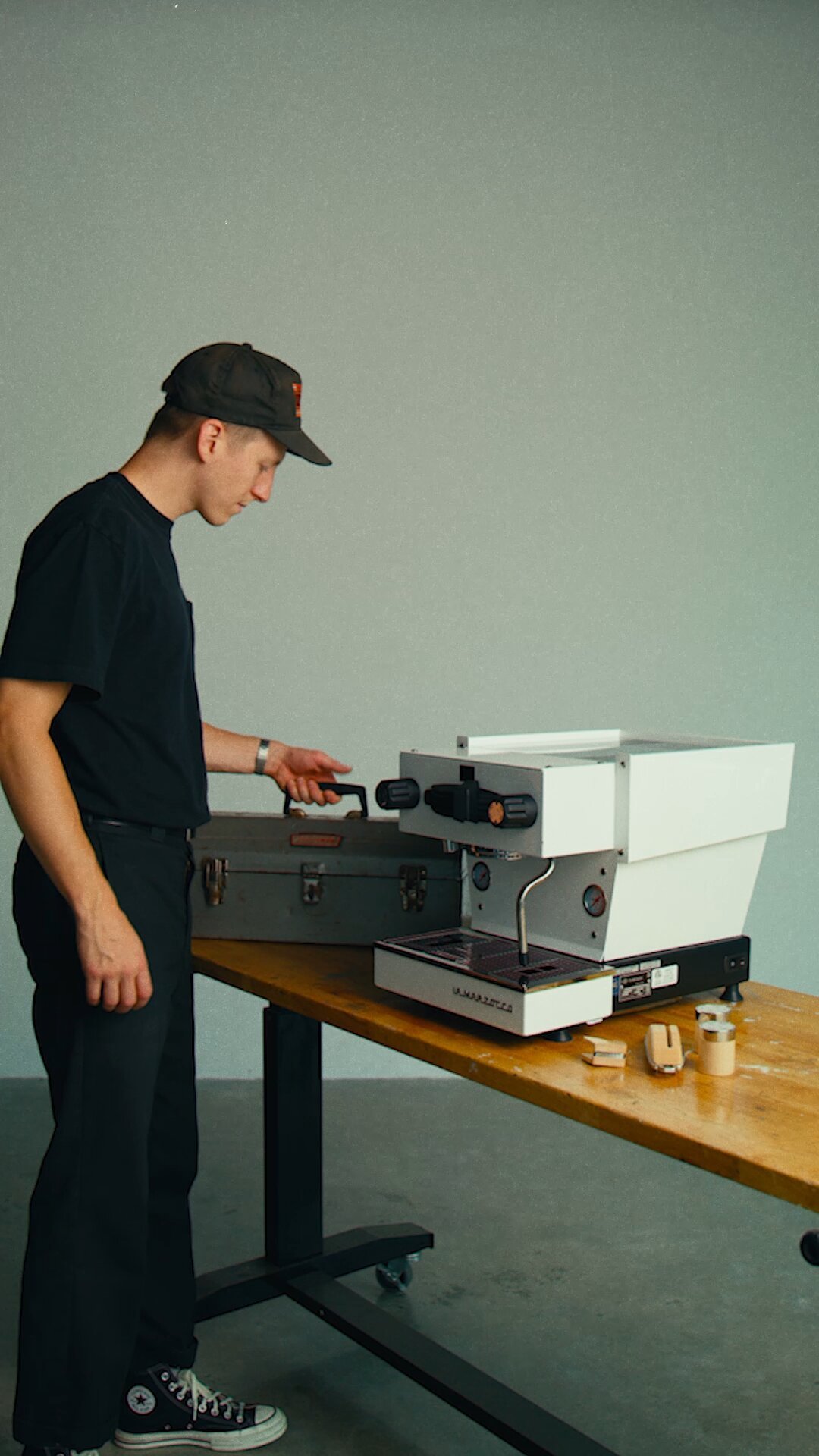 Person in black shirt and cap operating a La Marzocco Linea Mini espresso machine on a wooden table against a green wall