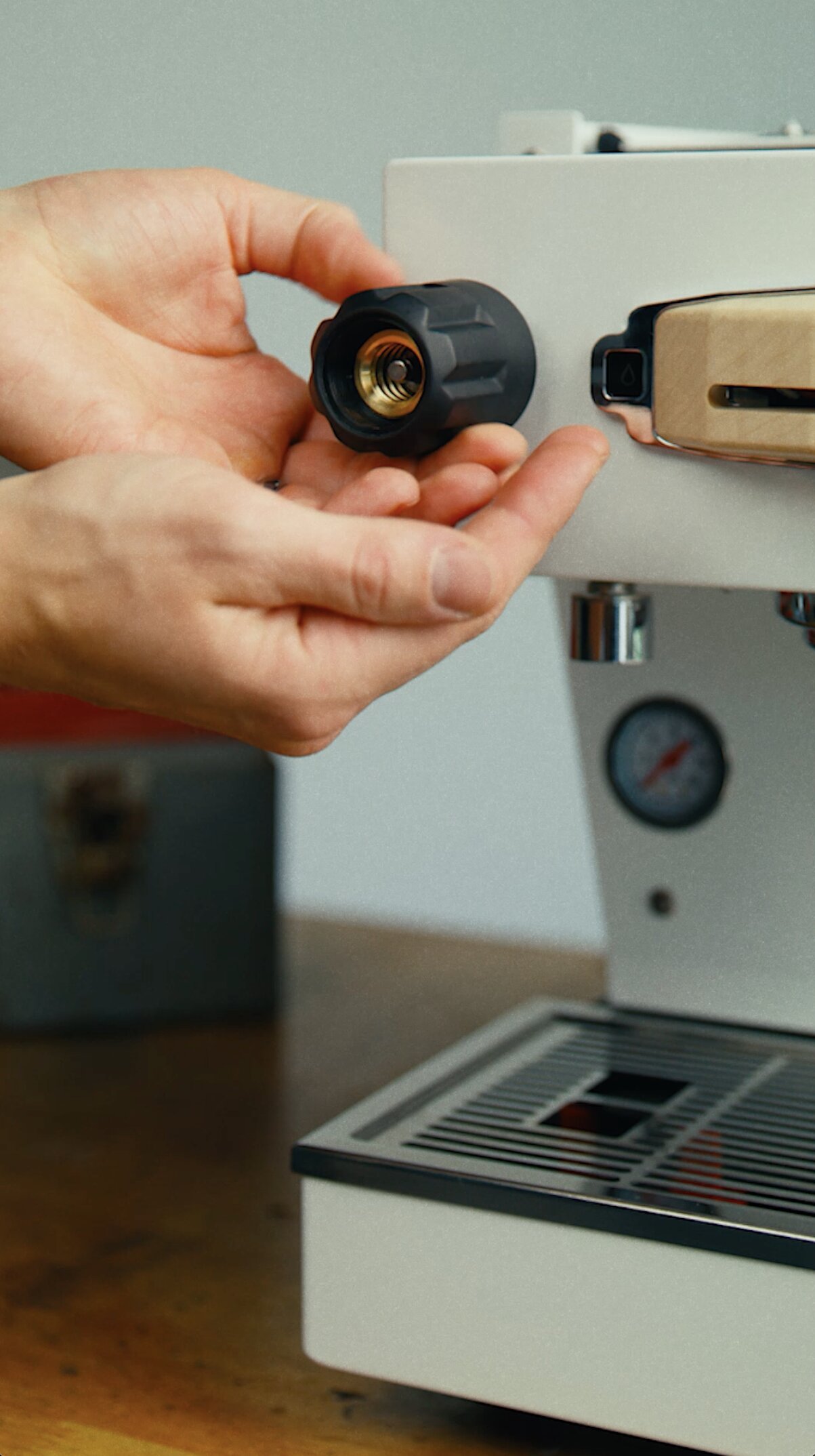 Hand holding La Marzocco group head connector with brass fitting, espresso machine in background