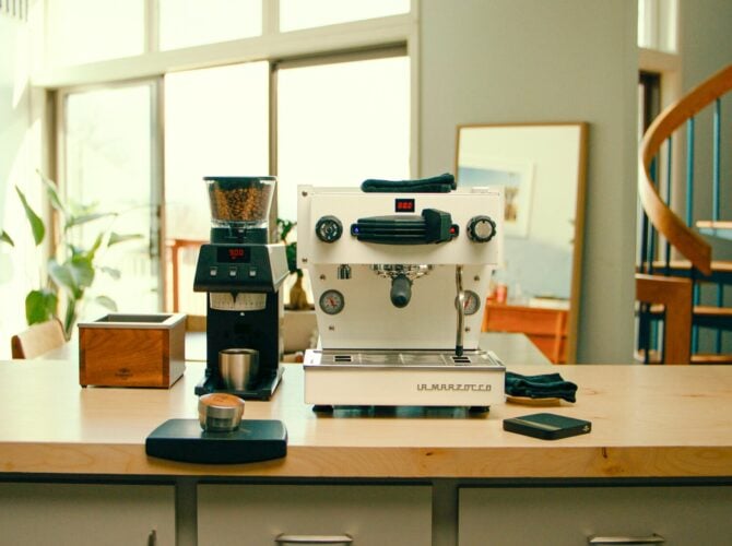 La Marzocco espresso machine and grinder displayed on a wooden counter in a bright home kitchen with plants and shelving