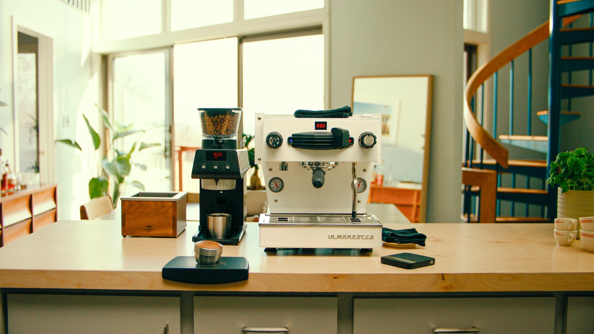 La Marzocco espresso machine and grinder displayed on a wooden counter in a bright home kitchen with plants and shelving