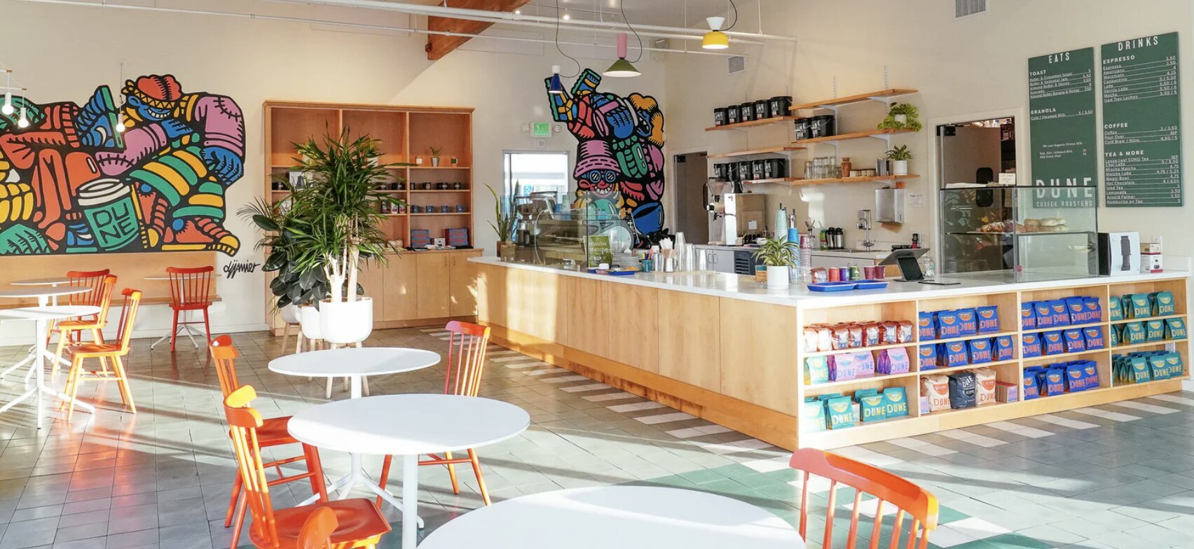 Modern café interior with La Marzocco espresso machines, colorful street art, wooden counter, and orange seating