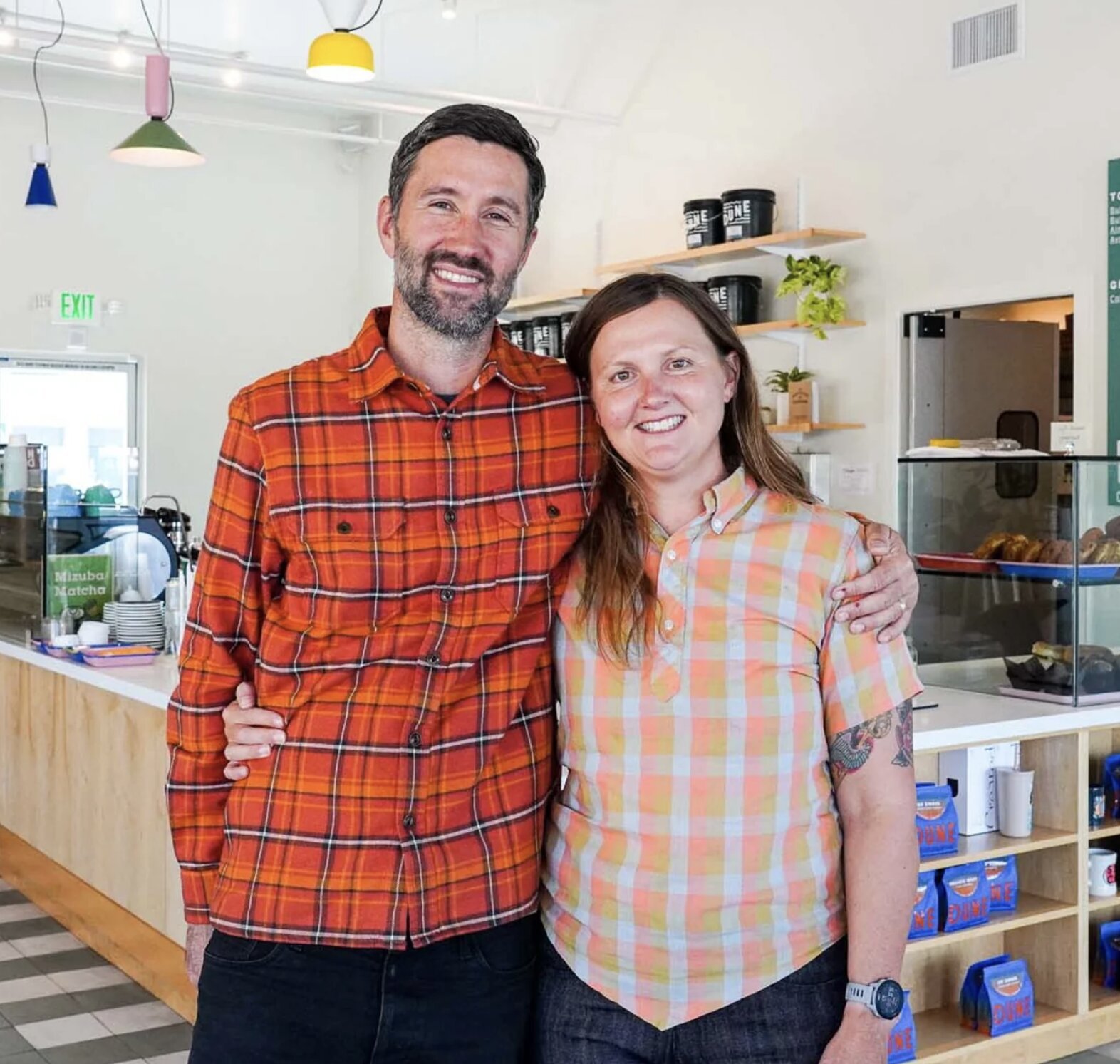 Two baristas smiling behind the counter of a modern coffee shop with La Marzocco espresso equipment and colorful pendant lighting