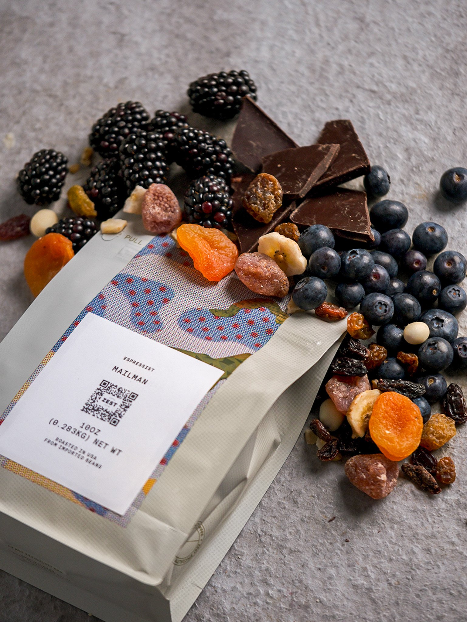Premium tea and chocolate tasting assortment with blackberries, blueberries, dried apricots, and dark chocolate shards on gray surface